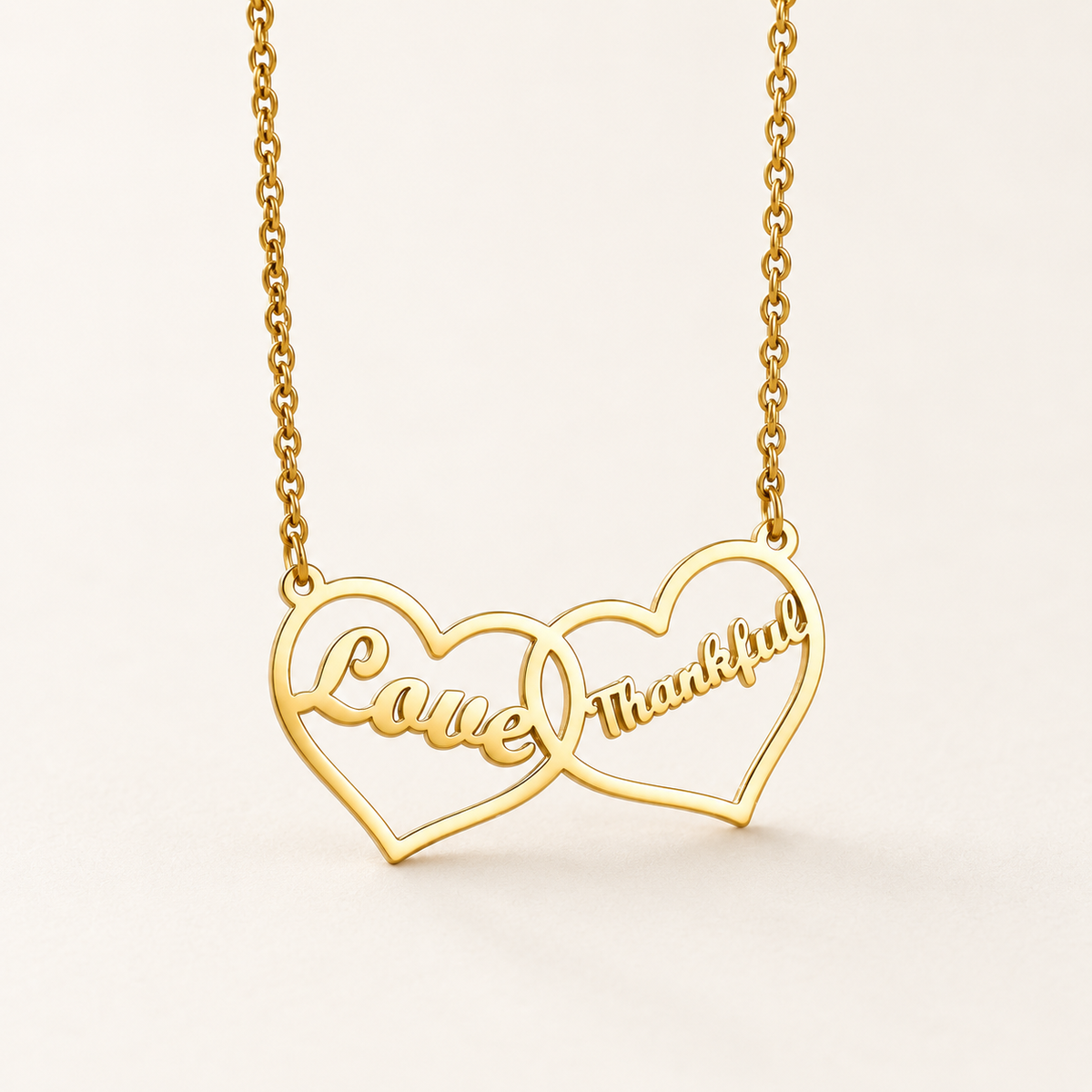 Goudkleurige ketting met twee verbonden hartjes en de voorbeeldgravures Love en Thankful op een rustige ivoorkleurige achtergrond