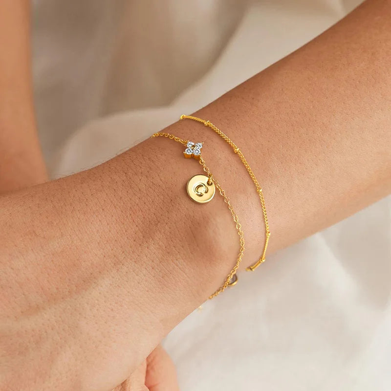 Dubbele goudkleurige armband met klavertje van zirkonia en lettermuntje om de pols