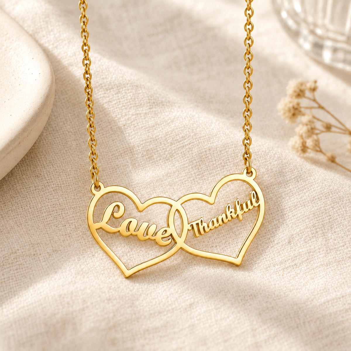 Goudkleurige ketting met twee verbonden hartjes en de voorbeeldgravures Love en Thankful op licht linnen in een warme setting