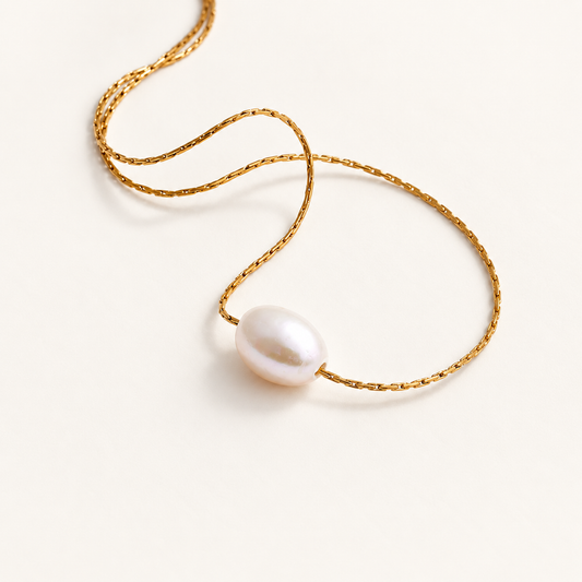 Goudkleurige ketting met één witte parel op een rustige ivoorkleurige achtergrond