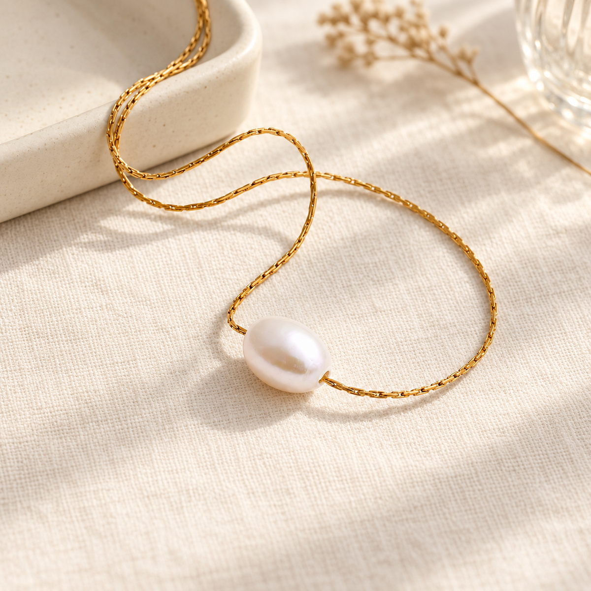 Goudkleurige ketting met één witte parel op licht linnen in een warme setting